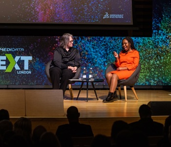 Two seated women on a stage talking in front of Medidata NEXT London signage