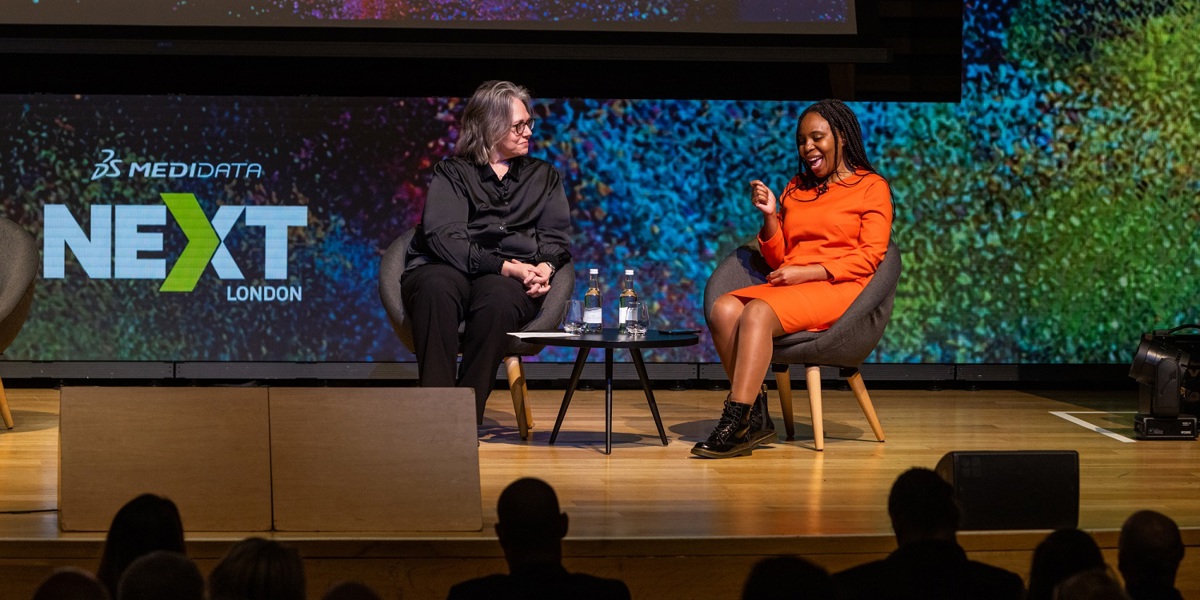 Two seated women on a stage talking in front of Medidata NEXT London signage