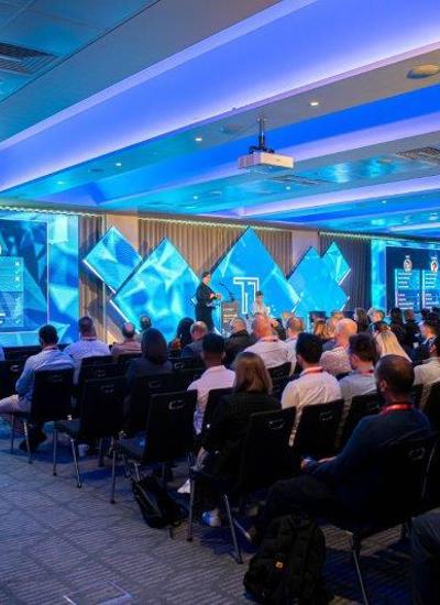 A room full of seated people watching a presentation given by a person on a stage