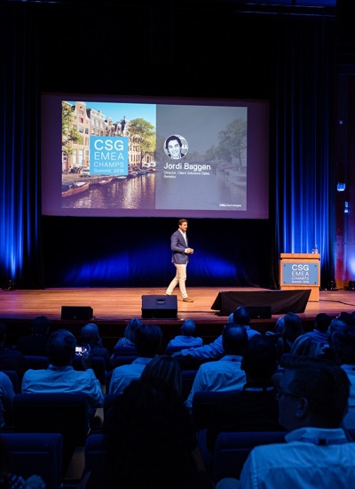 A room full of seated people watching a presentation given by a person on a stage